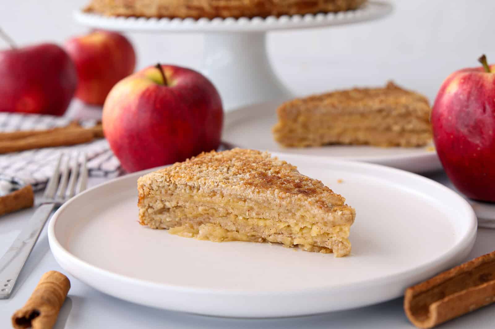 A slice of layered apple cake on a white plate, surrounded by red apples, cinnamon sticks, and a fork, with the rest of the cake and more apples in the background.