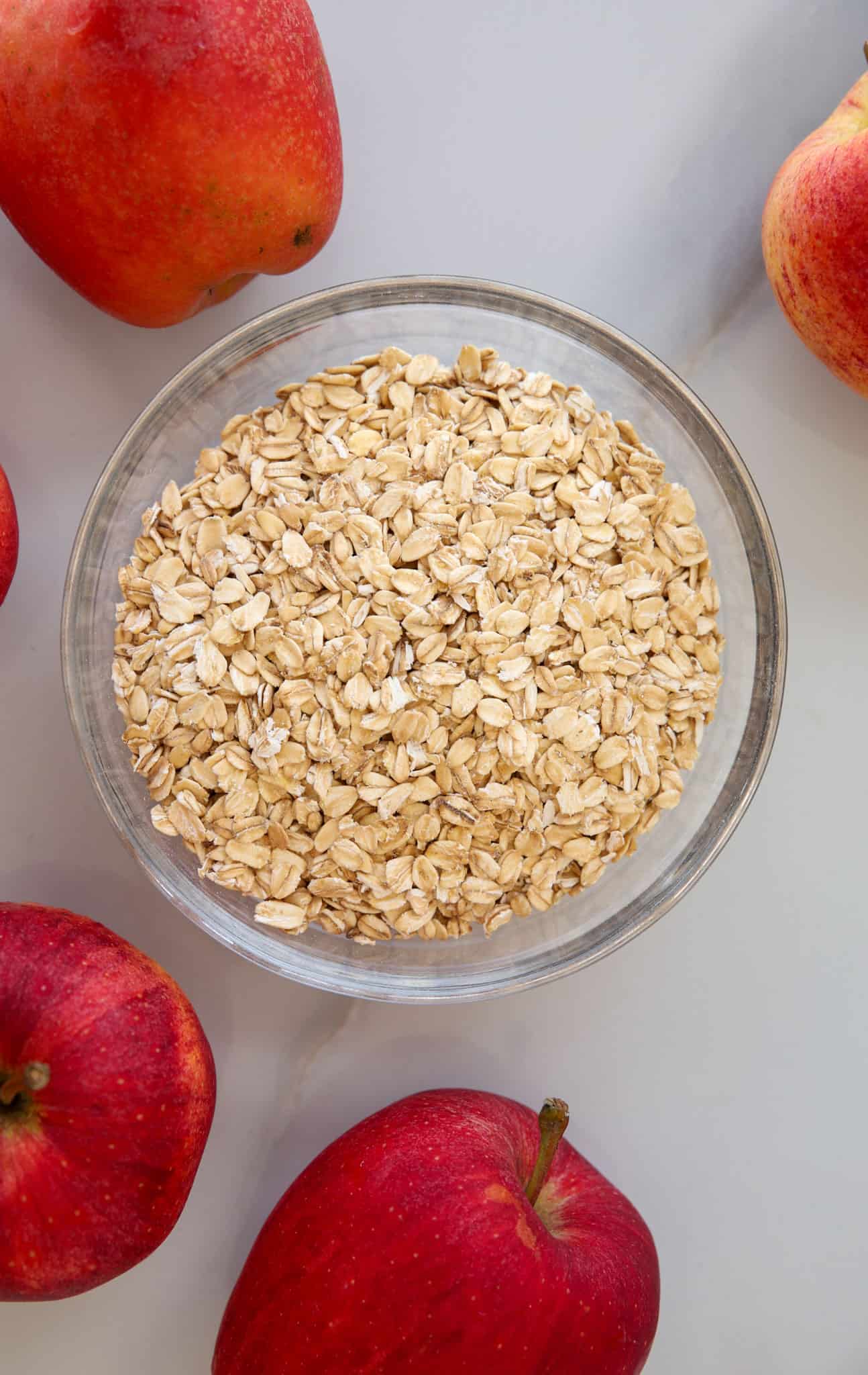 A clear glass bowl filled with rolled oats sits on a light surface, surrounded by several whole red apples.