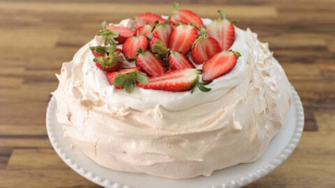 A pavlova topped with whipped cream and halved strawberries sits on a white cake stand. The crisp, light meringue has a slightly cracked surface, giving it a rustic appearance against a dark background.