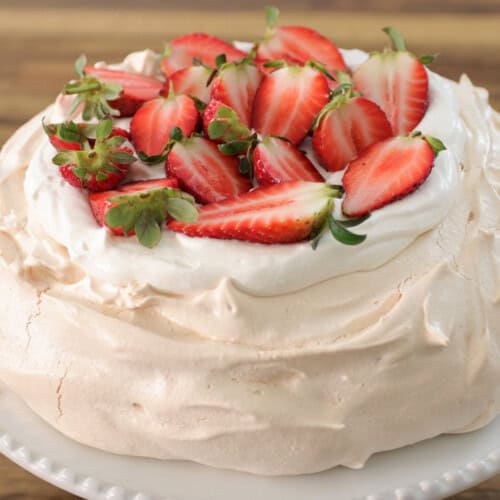 A pavlova topped with whipped cream and halved strawberries sits on a white cake stand. The crisp, light meringue has a slightly cracked surface, giving it a rustic appearance against a dark background.