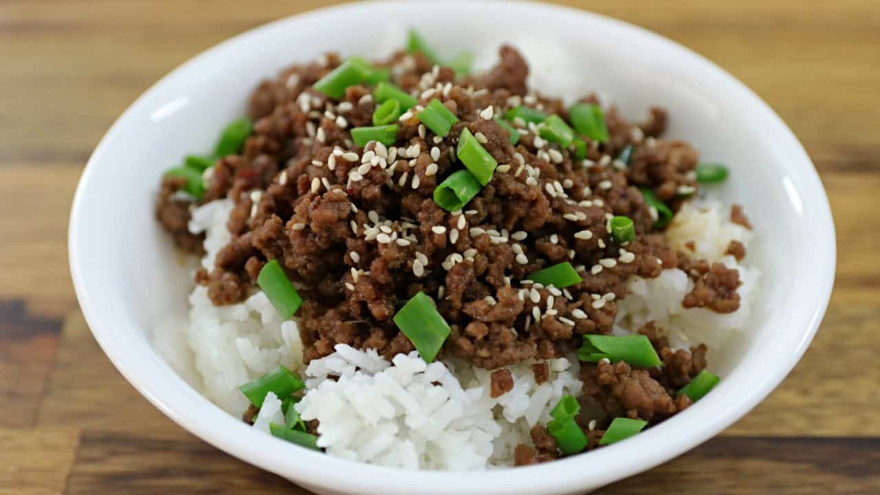 A white bowl filled with white rice topped with cooked ground beef, garnished with sesame seeds and chopped green onions. The bowl is set on a wooden surface.