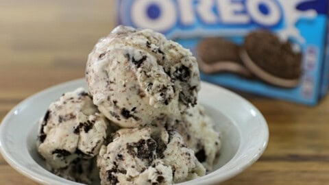 A close-up of a bowl filled with scoops of cookies and cream ice cream, featuring visible chunks of chocolate cookies. In the background, a partly visible Oreo cookie package is out of focus. The ice cream appears creamy and cold, perfect for a treat.