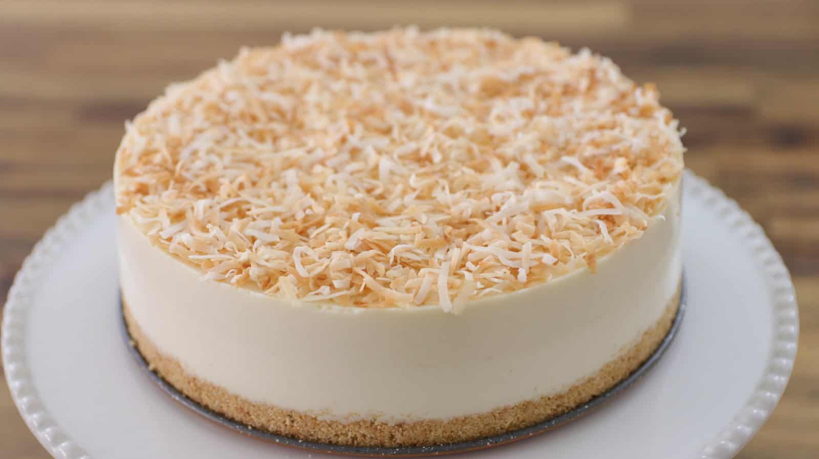 A hand is lifting a slice from a round coconut cheesecake. The cheesecake has a crumbly crust, a creamy filling, and is topped with toasted shredded coconut. It is served on a white plate with a textured edge, with a wooden surface in the background.