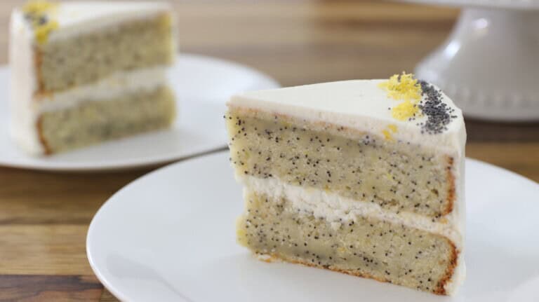 A whole lemon poppy seed cake with white frosting is topped with a lemon slice and garnished with poppy seeds and lemon zest. A slice of the cake is being lifted out with a metal server, showcasing the cake's interior. The background features a wooden surface.