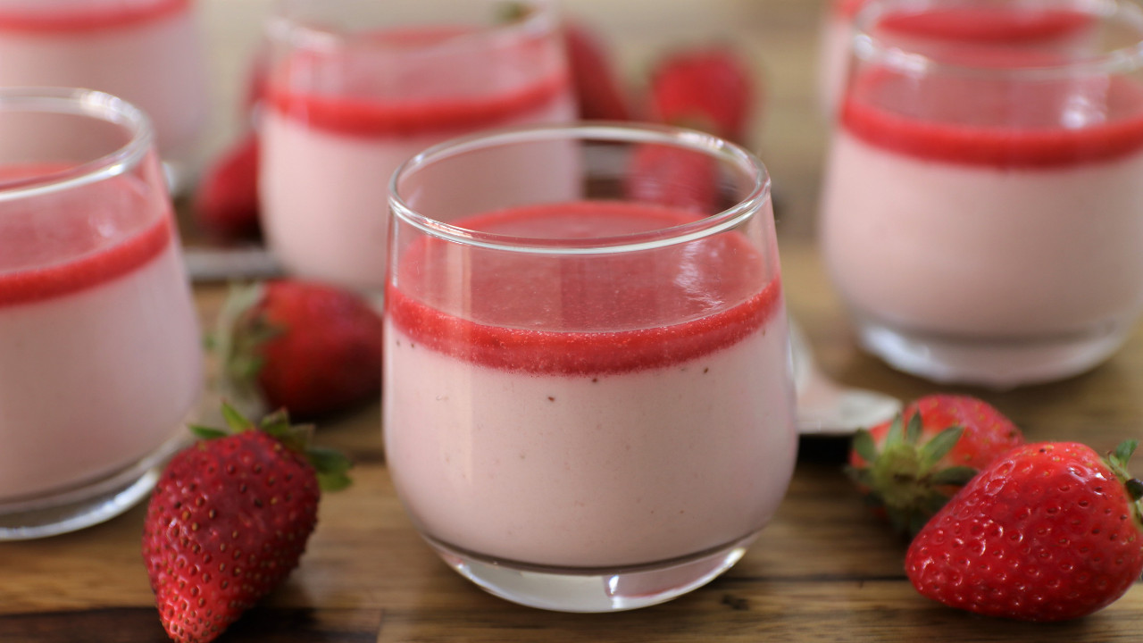 A glass of strawberry panna cotta topped with a layer of strawberry sauce is shown in the foreground. Additional glasses filled with mousse and several fresh strawberries are visible in the background, all placed on a wooden surface.