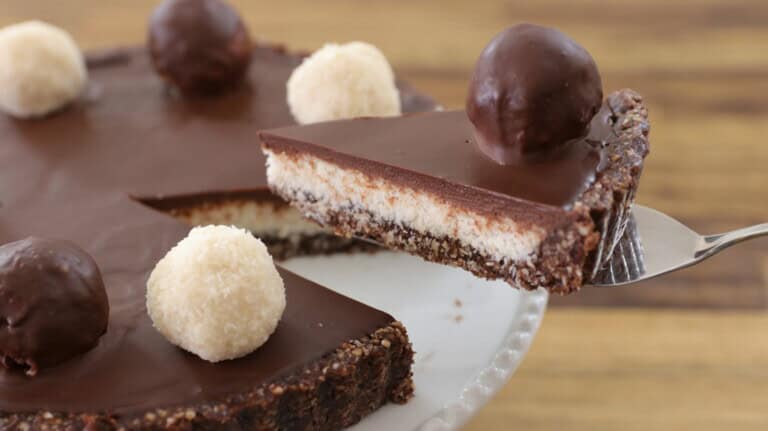 A close-up of a chocolate tart sits on a white plate. The tart features a smooth, glossy chocolate ganache filling, topped with round chocolate and white truffles. A slice is being lifted out with a serving utensil, revealing a layered crust and filling.