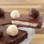 A close-up of a chocolate tart sits on a white plate. The tart features a smooth, glossy chocolate ganache filling, topped with round chocolate and white truffles. A slice is being lifted out with a serving utensil, revealing a layered crust and filling.