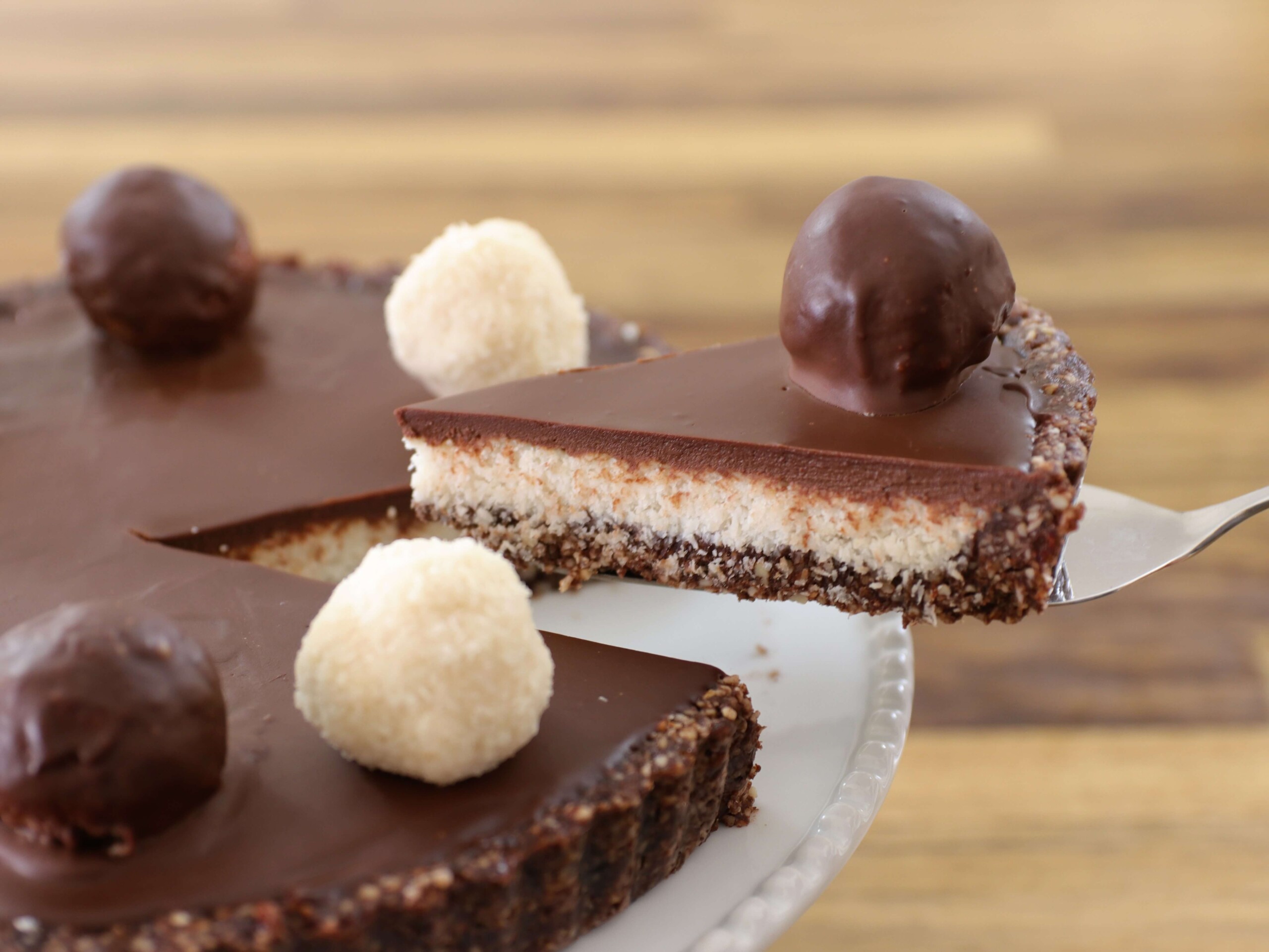 A close-up of a chocolate tart sits on a white plate. The tart features a smooth, glossy chocolate ganache filling, topped with round chocolate and white truffles. A slice is being lifted out with a serving utensil, revealing a layered crust and filling.