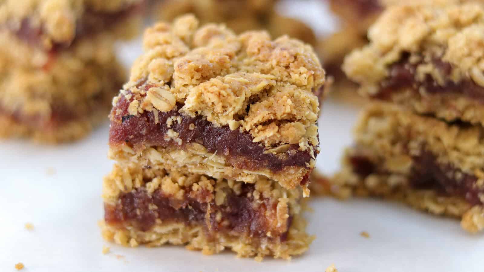 A close-up view of neatly arranged square date bars. Each bar has a crumbly oat topping and a dense, dark date filling in the middle, set on a wooden surface. The bars are aligned in multiple rows, creating a uniform pattern.