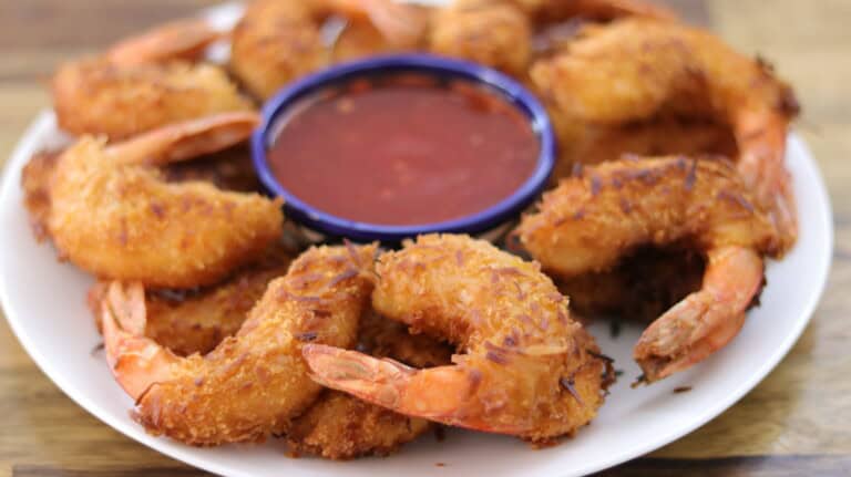 A plate of golden-brown fried shrimp arranged in a circle around a small bowl of red dipping sauce. The shrimp are crispy and have visible pieces of shredded coconut on the surface, indicating they are coconut shrimp. The bowl of sauce is placed at the center.