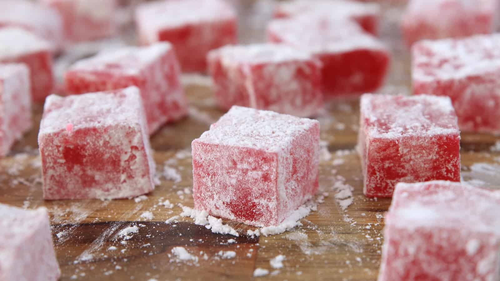 Close-up of several red Turkish delights dusted with powdered sugar, arranged on a blue plate. The cubes have a slightly translucent, chewy texture and are evenly coated with a fine layer of sugar, giving them a frosty appearance.