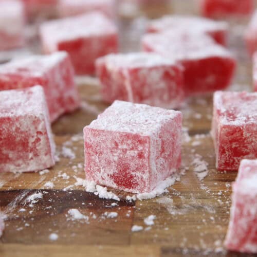 Close-up of several red Turkish delights dusted with powdered sugar, arranged on a blue plate. The cubes have a slightly translucent, chewy texture and are evenly coated with a fine layer of sugar, giving them a frosty appearance.