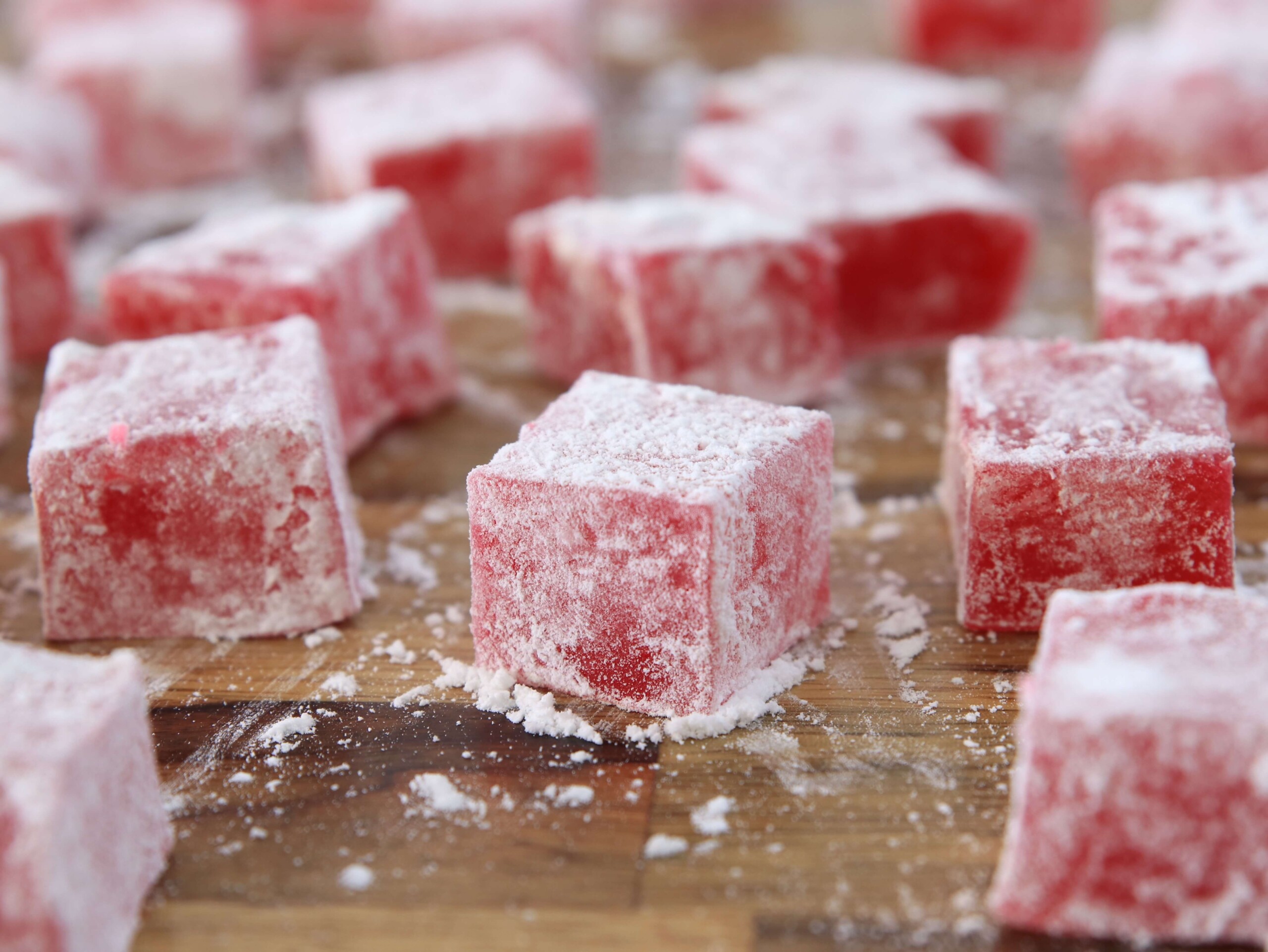 Close-up of several red Turkish delights dusted with powdered sugar, arranged on a blue plate. The cubes have a slightly translucent, chewy texture and are evenly coated with a fine layer of sugar, giving them a frosty appearance.