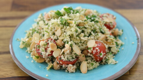 A vibrant plate of cauliflower rice salad is served. The dish includes finely chopped cauliflower, halved cherry tomatoes, chickpeas, and green herbs, all topped with sliced almonds. The ingredients are fresh, colorful, and arranged on a blue plate.