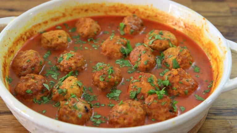 Round chicken meatballs in a rich red coconut curry sauce, garnished with chopped fresh herbs, served in a white dish on a wooden table.