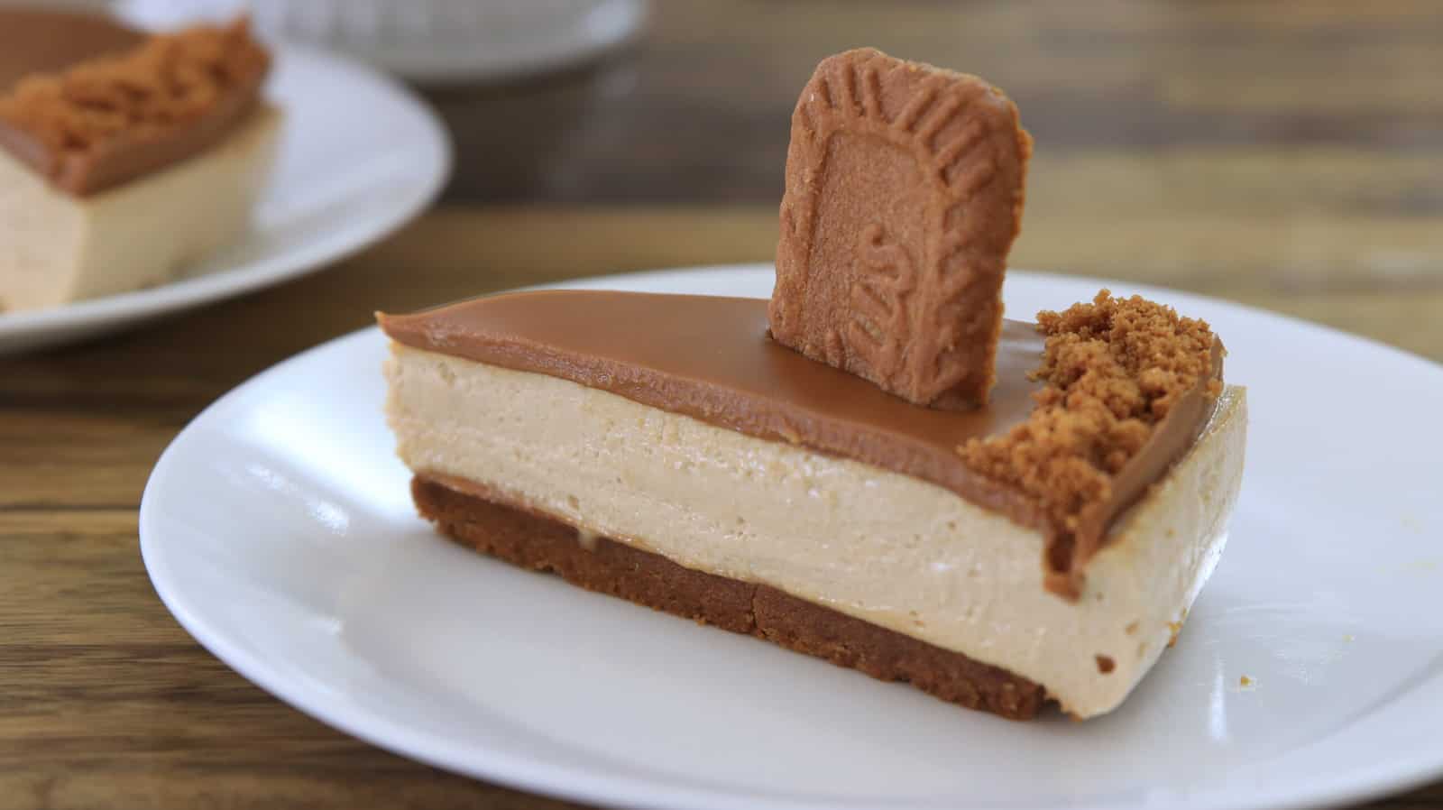 A creamy biscoff cheesecake sits on a white fluted cake stand. The cheesecake has a smooth caramel topping and a crumbly crust around the outer edge. The background is a wooden surface.