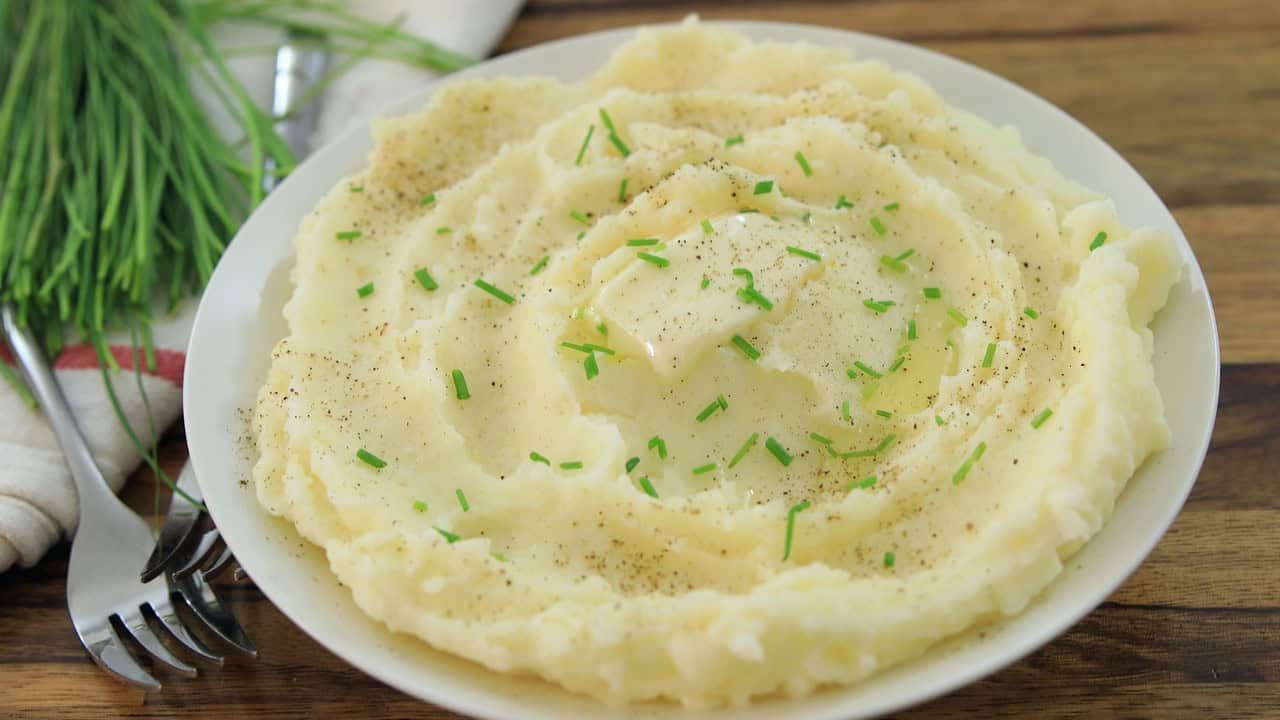 A white plate filled with creamy mashed potatoes garnished with chopped chives and a sprinkle of black pepper, served on a wooden table with a fork and napkin beside it. Fresh green chives are visible in the background.