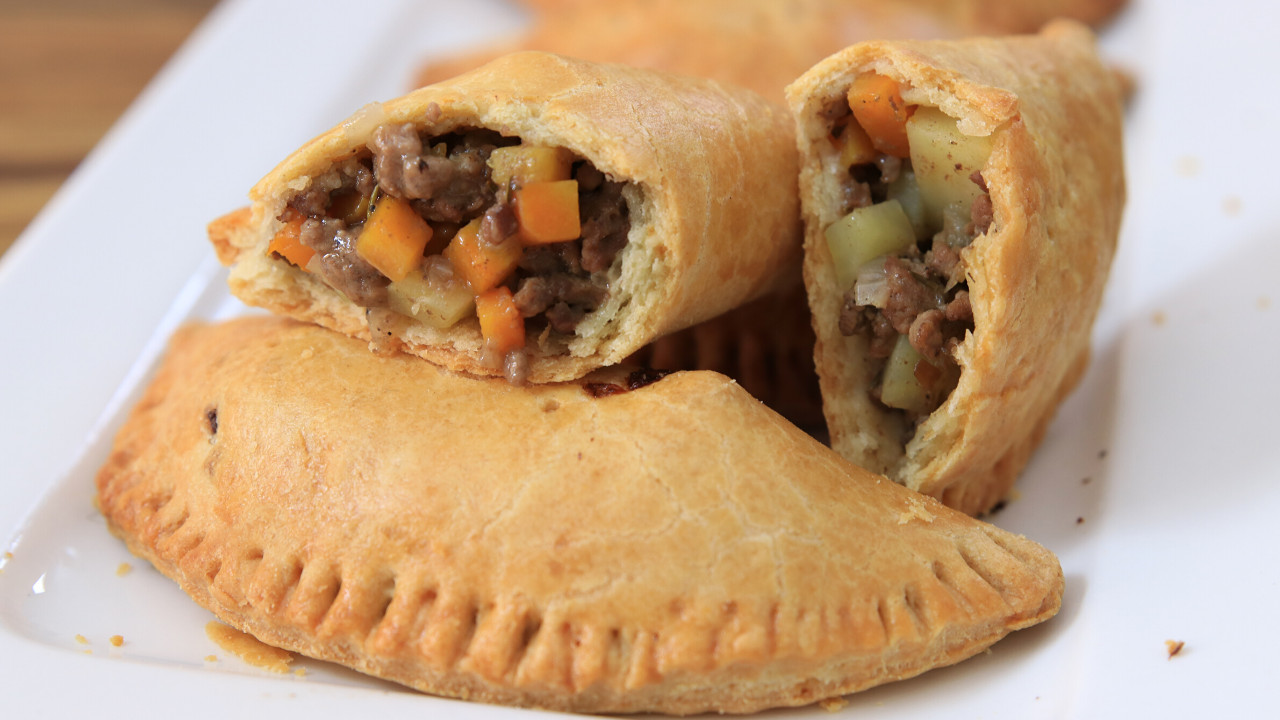 A plate holds two empanadas, one whole and one cut in half. The crispy golden-brown pastry reveals a filling of diced vegetables and meat. The filling appears to contain beef, carrots, and potatoes, showcasing a savory and colorful combination.