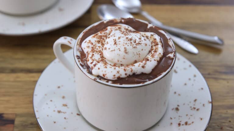 A white ceramic mug filled with hot chocolate, topped with a swirl of whipped cream and sprinkled with chocolate shavings. The mug sits on a matching saucer with chocolate shavings around it. In the background, there is another saucer with two spoons.