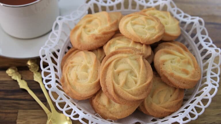 A basket filled with golden, swirl-patterned butter cookies is placed on a wooden table. The basket is lined with a white doily. In the background, there is a white teacup and saucer, and a couple of ornate gold spoons are resting beside the basket.