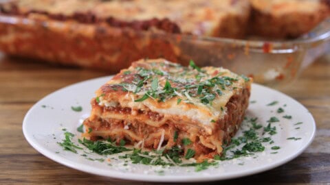 A close-up of a slice of lasagna on a white plate. The lasagna has visible layers of pasta, meat sauce, and cheese, topped with finely chopped parsley and grated Parmesan. In the background, a baking dish with the remaining lasagna is partially visible.