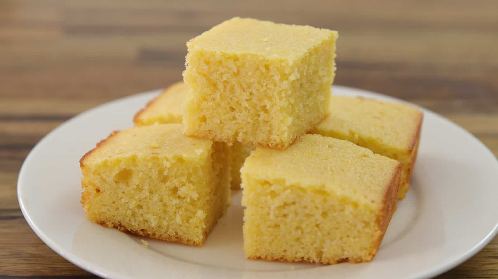 Five pieces of freshly baked, golden cornbread stacked on a white plate against a wooden background. The cornbread squares have a moist and fluffy texture with slightly browned edges.