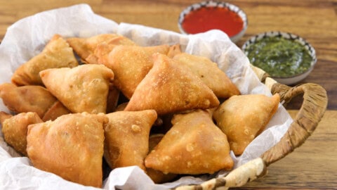 A woven basket lined with white paper holds several golden-brown, triangular samosas. In the background, two small bowls of dipping sauces are visible, one green and one red. The scene is set on a wooden surface.