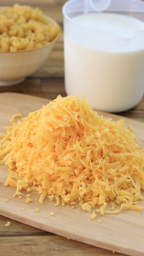 A pile of shredded cheddar cheese is on a wooden cutting board, with a container of uncooked macaroni and a large measuring cup of milk in the background. The scene appears ready for making mac and cheese.