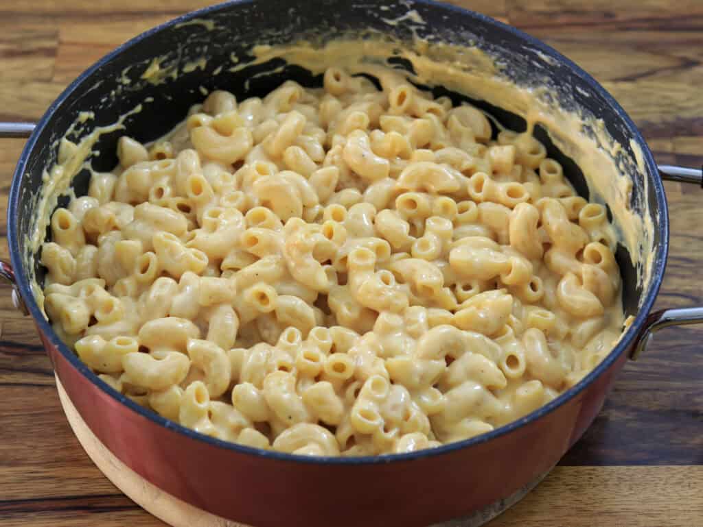 A large, orange-hued pot filled with creamy macaroni and cheese. The macaroni noodles are coated in a rich, melted cheese sauce. The pot rests on a wooden surface.