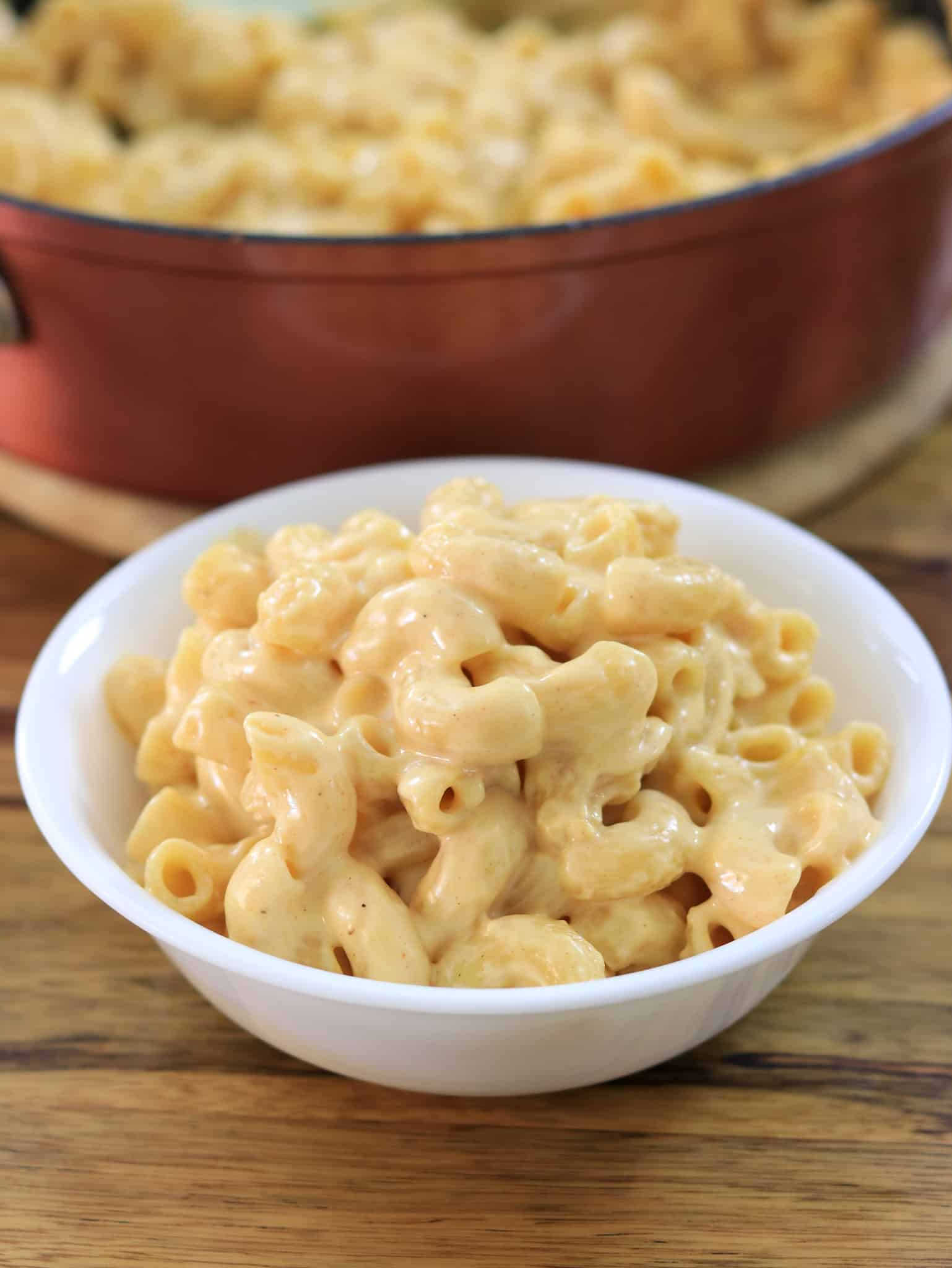 A white bowl filled with creamy macaroni and cheese is placed on a wooden table. In the background, a larger serving of macaroni and cheese in a red pot is partially visible.