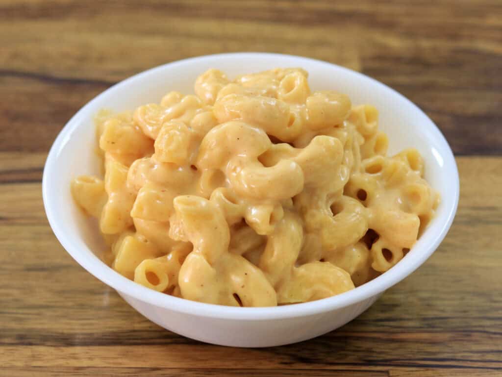 A white bowl filled with creamy macaroni and cheese sits on a wooden surface. The pasta is generously coated in a smooth, yellow cheese sauce.