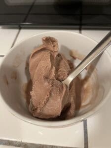 A white bowl with chocolate ice cream and a silver spoon, sitting on a tiled countertop.