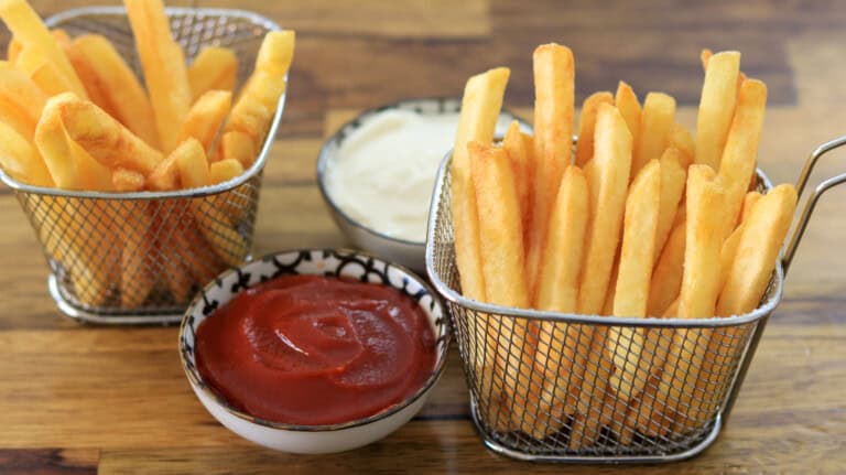 Two metal baskets filled with golden, crispy French fries are placed on a wooden table. Between them, there are two small bowls, one filled with ketchup and the other with a creamy white sauce.