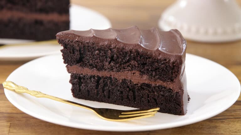 Two slices of rich chocolate cake with thick, glossy chocolate frosting are placed on white plates. Each plate includes a golden fork, and the chocolate cake layers appear moist and decadent. The background is a light wooden surface.