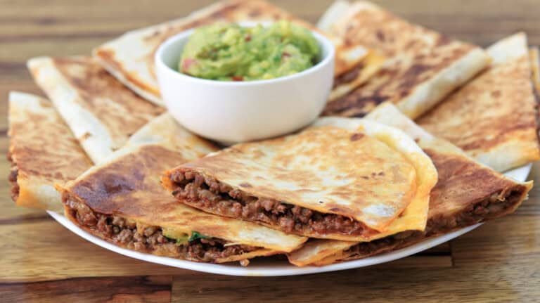 A plate of golden-brown quesadillas, filled with seasoned ground beef, sits on a wooden surface. The quesadillas are neatly arranged around a small white bowl of guacamole placed in the center of the plate.