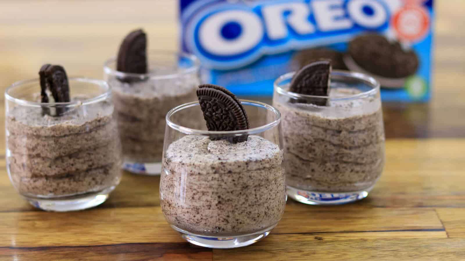 Four glasses filled with cookies and cream mousse are neatly arranged in a row on a wooden surface. Each glass is topped with a half Oreo cookie. In the background, there is a blurred Oreo cookie package.