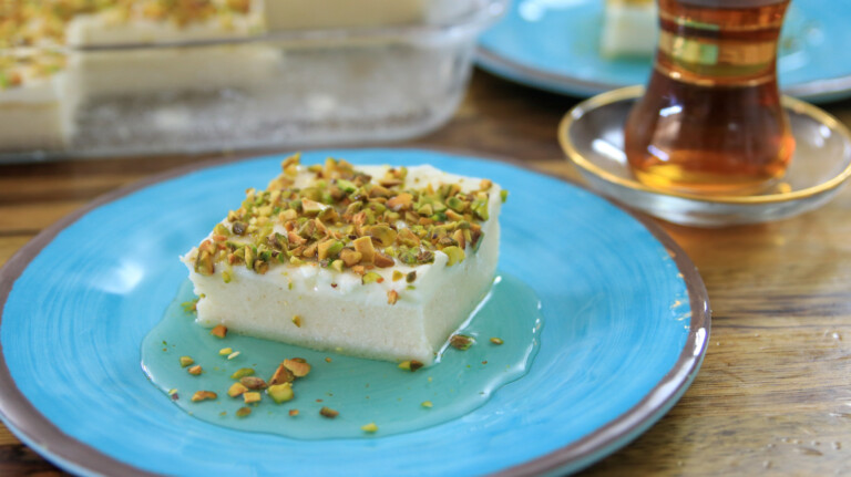 A slice of white dessert topped with chopped pistachios sits on a blue plate. There is a small golden dessert fork next to it on the plate. In the background, a glass of tea on a saucer is visible.