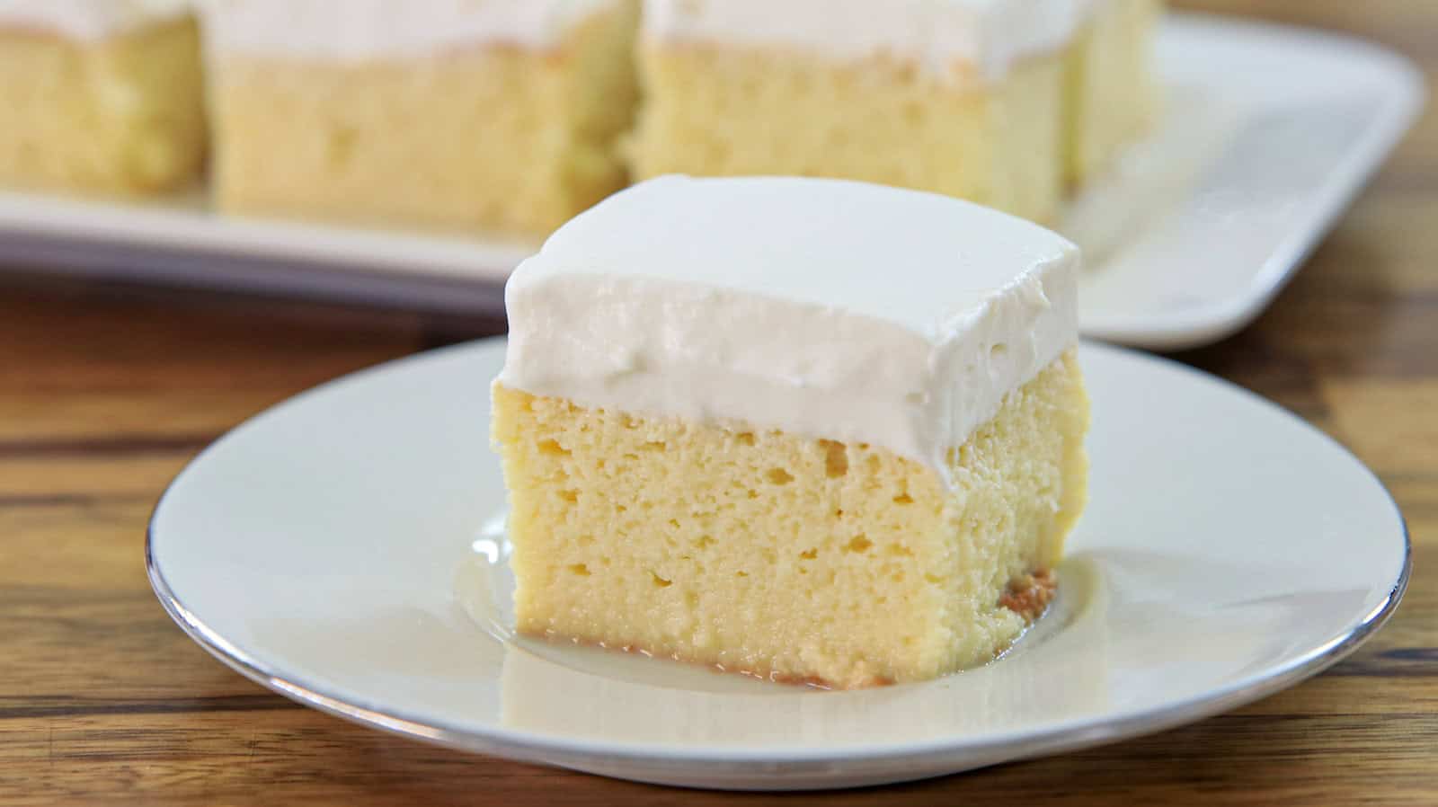 A slice of tres leches cake sits on a white plate. The cake is topped with a layer of fluffy white frosting, and the moist, pale yellow base is visible. In the background, more slices of cake are displayed on a rectangular serving dish.