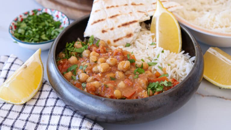 A bowl of chickpea stew with tomatoes and herbs, served with white rice, grilled flatbread, and lemon wedges. Chopped herbs and extra lemon are on the side, with a checkered napkin nearby.