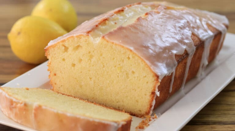 A loaf of lemon pound cake with a slice cut off is placed on a white rectangular plate. The cake has a thick glaze on top and two whole lemons are visible in the background, all set on a wooden surface.