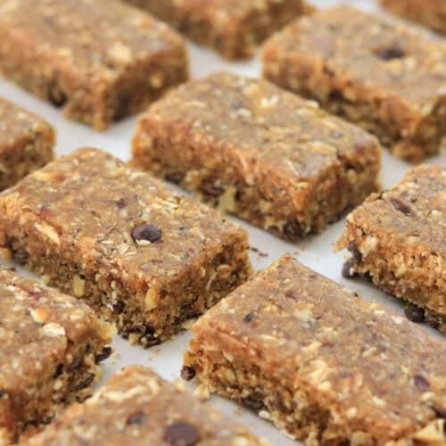 Rows of homemade granola bars, featuring a mixed texture with visible oats, nuts, and chocolate chips, placed on parchment paper. The bars have a natural, rustic appearance, arranged neatly in a grid pattern.