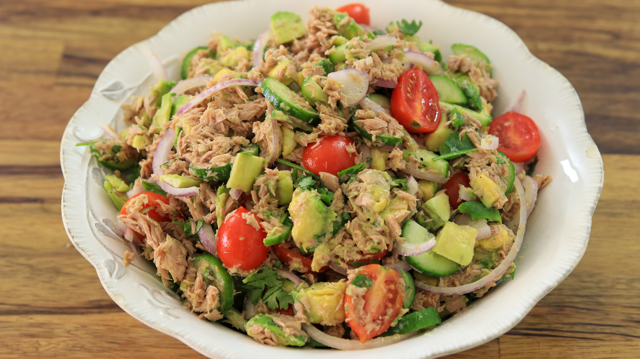 A large, white bowl filled with a colorful salad. The salad has chunks of tuna, sliced red onions, halved cherry tomatoes, diced avocados, cucumber slices, and fresh cilantro. The bowl is placed on a wooden surface.