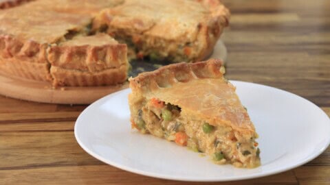 A slice of chicken pot pie is served on a white plate in the foreground, with the rest of the pie on a wooden board in the background. The pie has a golden-brown crust, and the filling appears to contain chicken and mixed vegetables. The text "CHICKEN POT PIE" is prominently displayed.