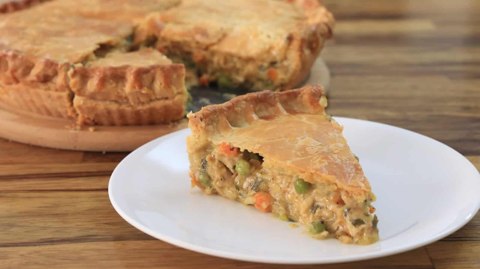 A slice of chicken pot pie is served on a white plate in the foreground, with the rest of the pie on a wooden board in the background. The pie has a golden-brown crust, and the filling appears to contain chicken and mixed vegetables. The text "CHICKEN POT PIE" is prominently displayed.