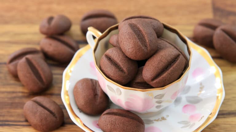 A teacup filled with several chocolate coffee bean-shaped cookies sits on a matching saucer. More cookies are scattered on the wooden table around the cup. The cookies are dark brown with a central line, resembling coffee beans.