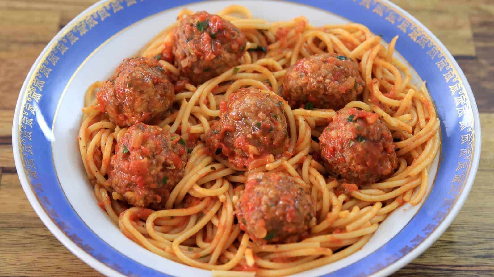 A dish of spaghetti and meatballs in tomato sauce is served on a plate with a blue and white rim. The vibrant red sauce coats the pasta and meatballs, and the dish is placed on a wooden surface.