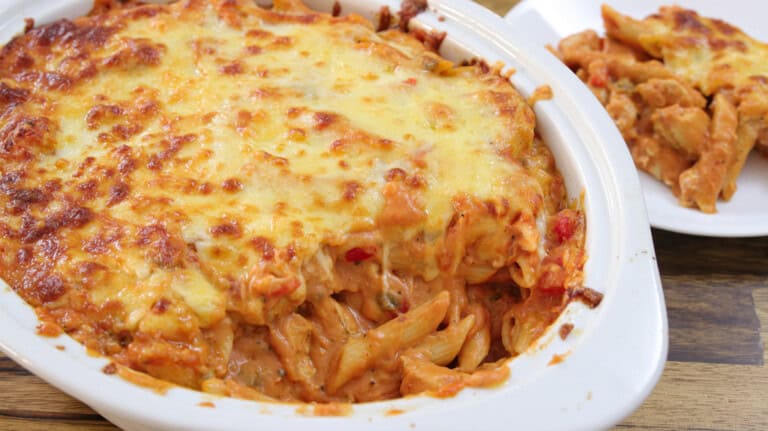 A close-up of a baked pasta dish with melted cheese on top in a white casserole dish. A portion has been scooped out, revealing creamy pasta with red sauce and visible chunks of tomato and herbs inside. The dish rests on a wooden surface.