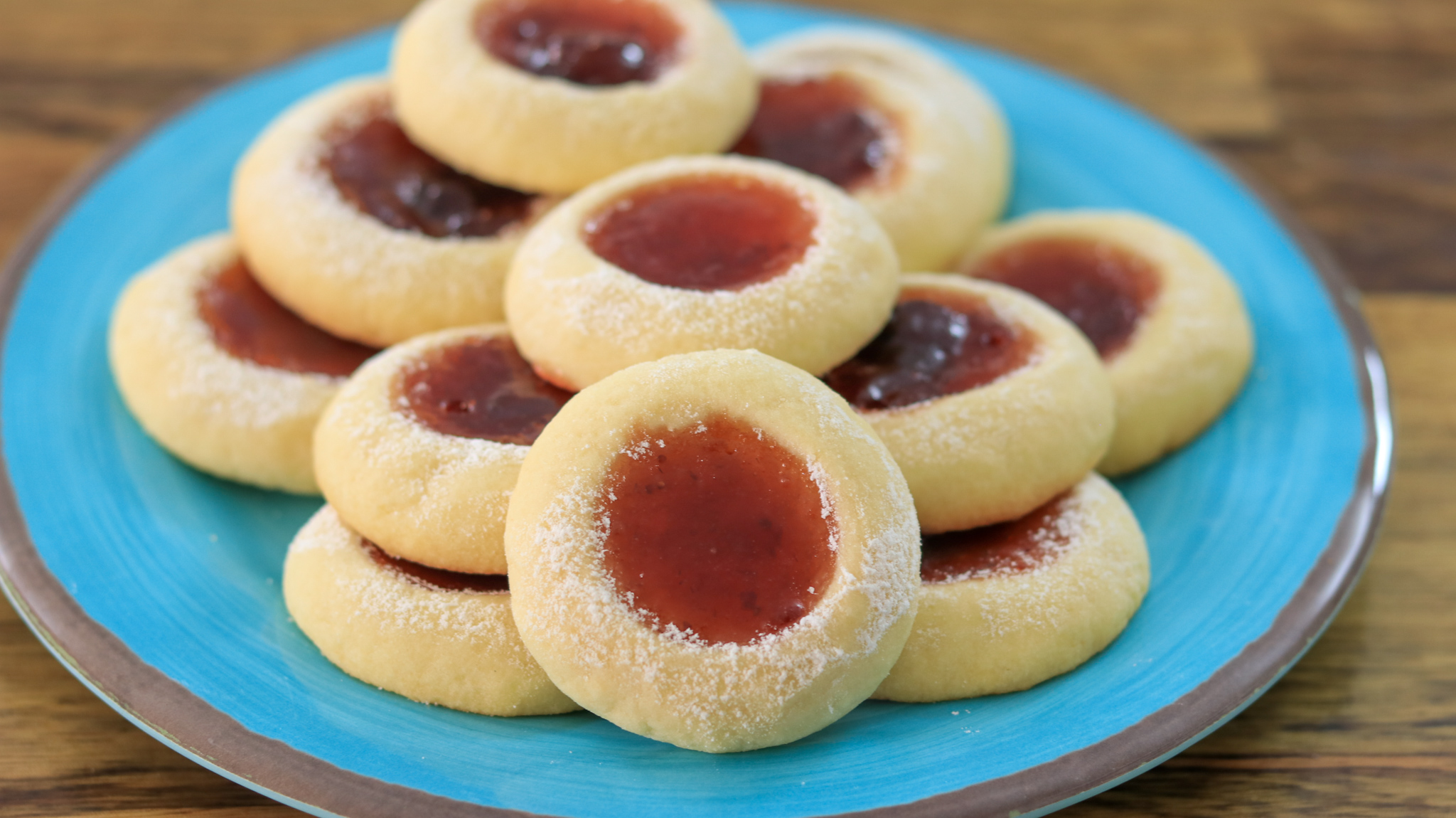 A cooling rack holds several jam-filled thumbprint cookies. The cookies have a light dusting of powdered sugar and are filled with different jams, with some centers appearing red and others a darker purple. The wooden surface underneath adds a rustic feel.