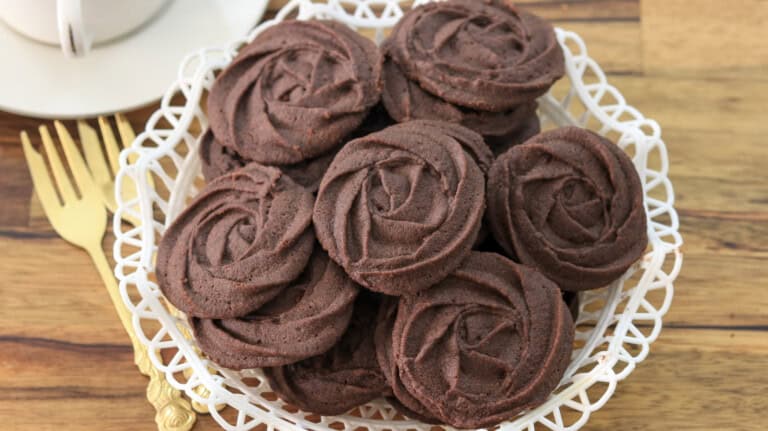 A white lace-patterned plate holds an arrangement of chocolate cookies, each shaped like a rose. The plate is placed on a wooden surface next to a gold fork and a white teacup and saucer.