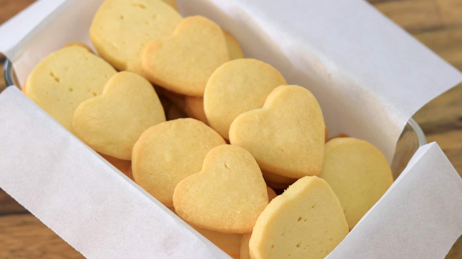 A tray lined with white parchment paper holds several shortbread cookies. Some of the cookies are heart-shaped, while others are round. The cookies have a uniform, golden-brown color, indicating they are freshly baked.